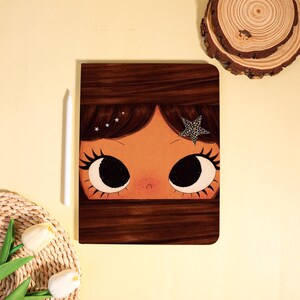 May include: A brown notebook with an illustration of a girl's face, featuring large black eyes, long eyelashes, and a star-shaped hair accessory. A white stylus rests beside the notebook on a yellow surface.