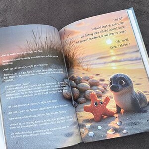 以下が含まれることがあります： 浜辺にいるアザラシとヒトデのイラストが描かれた、開かれた子供向けの本。本のページにはカラフルな画像とドイツ語のテキストが掲載されています。表紙は水色で、背景は濃いグレーです。