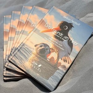 以下が含まれることがあります： 「Drei Freunde am Meer」というタイトルの子供向けの本が複数冊。表紙には鳥とラッコが描かれています。本は砂の表面に置かれています。