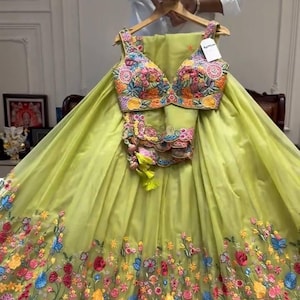 Può includere: Un completo lehenga choli verde lime ricamato. La gonna e il top presentano ricami floreali colorati. Il top ha uno scollo a V profondo e la gonna è lunga fino al pavimento. Il completo è esposto su una gruccia di legno.