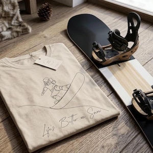 Op de afbeelding: Een beige t-shirt met een lijntekening van een snowboarder en de tekst "Life's Better on Snow". Een zwart-wit snowboard met bindingen ligt naast het shirt. Het shirt heeft een label.