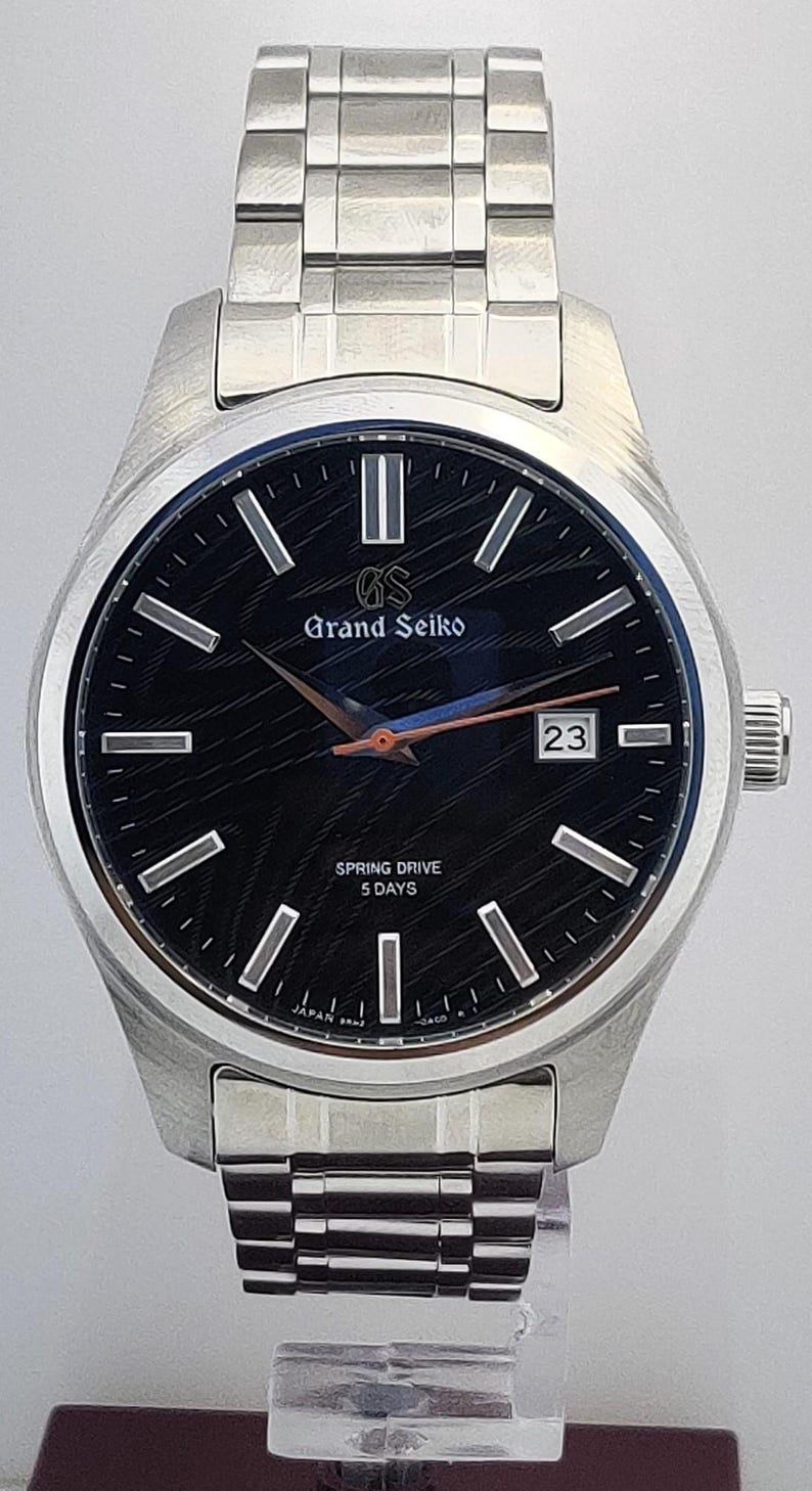 K&ouml;nnte beinhalten: Eine Grand Seiko-Uhr aus Edelstahl mit schwarzem Zifferblatt und silbernen Zeigern. Das Zifferblatt zeigt den Text "Grand Seiko" und "Spring Drive 5 Days". Das Datum wird auf 3 Uhr angezeigt.