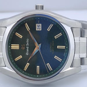 May include: A stainless steel wristwatch with a dark green dial and gold-colored hands and markers. The watch face displays the date and the words "Grand Seiko" and "Automatic Hi-Beat 36000". The watch band is made of stainless steel.