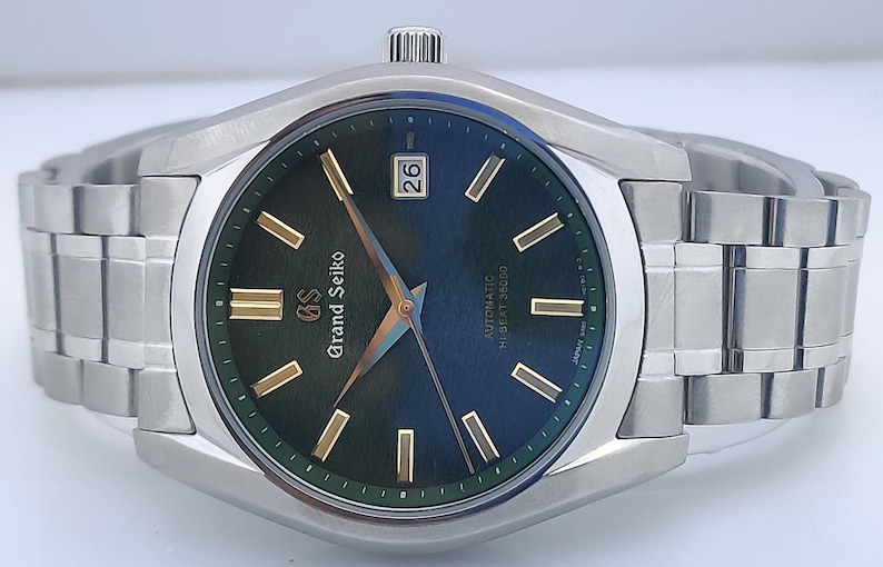 May include: A stainless steel wristwatch with a dark green dial and gold-colored hands and markers. The watch face displays the date and the words "Grand Seiko" and "Automatic Hi-Beat 36000". The watch band is made of stainless steel.