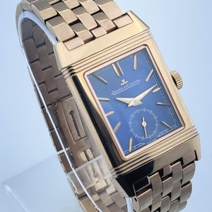 May include: A gold-toned rectangular wristwatch with a blue face. The watch features a gold band with a woven design. The watch face has gold hands and the brand name "Jaeger-LeCoultre".