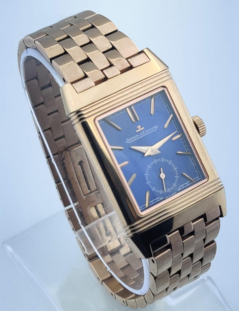 May include: A gold-toned rectangular wristwatch with a blue face. The watch features a gold band with a woven design. The watch face has gold hands and the brand name "Jaeger-LeCoultre".