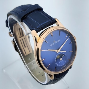 May include: A luxury wristwatch with a blue face and gold-colored casing. The watch features a dark blue leather band and the brand name "JAEGER-LECOULTRE" is visible on the face. The watch has a moon phase indicator.