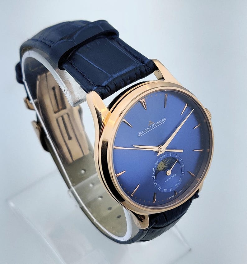 May include: A luxury wristwatch with a blue face and gold-colored casing. The watch features a dark blue leather band and the brand name "JAEGER-LECOULTRE" is visible on the face. The watch has a moon phase indicator.
