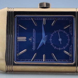 May include: A gold-toned rectangular wristwatch with a navy blue face. The watch features a small seconds dial and gold-toned hands and markers. The watch band is made of gold-toned rectangular links.