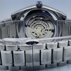 May include: Close-up of a silver-toned wristwatch with a metal band. The watch face is visible, showcasing the intricate inner workings and a gold-colored emblem. The band is composed of rectangular links.