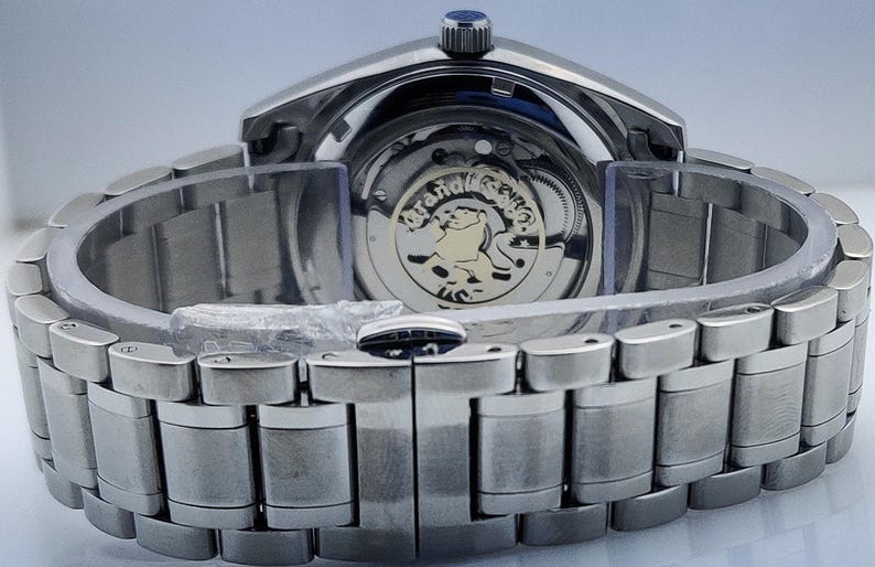 May include: Close-up of a silver-toned wristwatch with a metal band. The watch face is visible, showcasing the intricate inner workings and a gold-colored emblem. The band is composed of rectangular links.