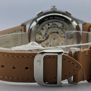 May include: Close-up of a luxury wristwatch with a light brown leather strap. The watch has a silver-colored case and a visible mechanical movement. The strap is secured with a silver buckle. The watch is a high-end accessory.