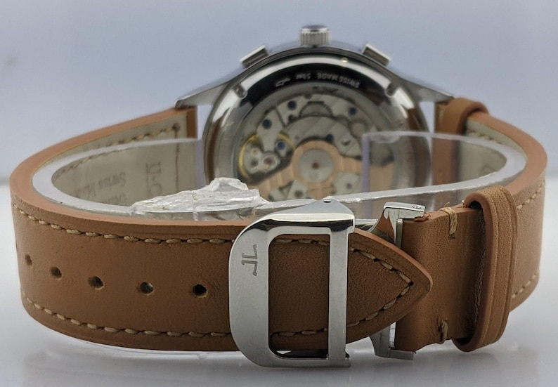 May include: Close-up of a luxury wristwatch with a light brown leather strap. The watch has a silver-colored case and a visible mechanical movement. The strap is secured with a silver buckle. The watch is a high-end accessory.