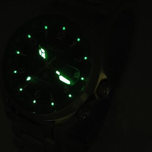 May include: Close-up of a Seiko automatic watch with a dark green face and luminous green markers and hands. The watch face displays the Seiko logo and the word "Automatic". The watch is set against a dark background.