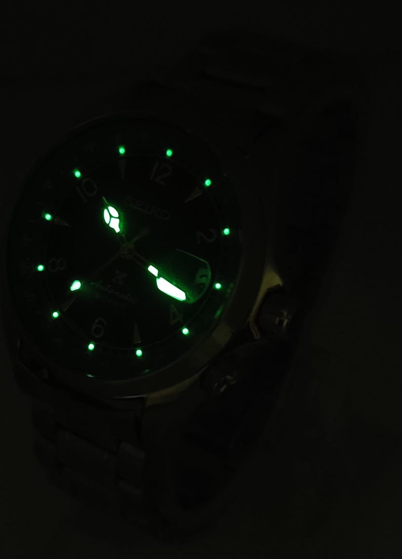 May include: Close-up of a Seiko automatic watch with a dark green face and luminous green markers and hands. The watch face displays the Seiko logo and the word "Automatic". The watch is set against a dark background.