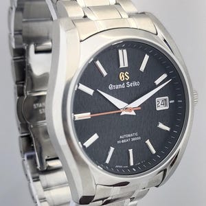 May include: A stainless steel Grand Seiko automatic wristwatch with a black dial. The watch features silver hands and markers, a date window, and the text "Grand Seiko" and "AUTOMATIC HI-BEAT 36000". The watch band is also stainless steel.