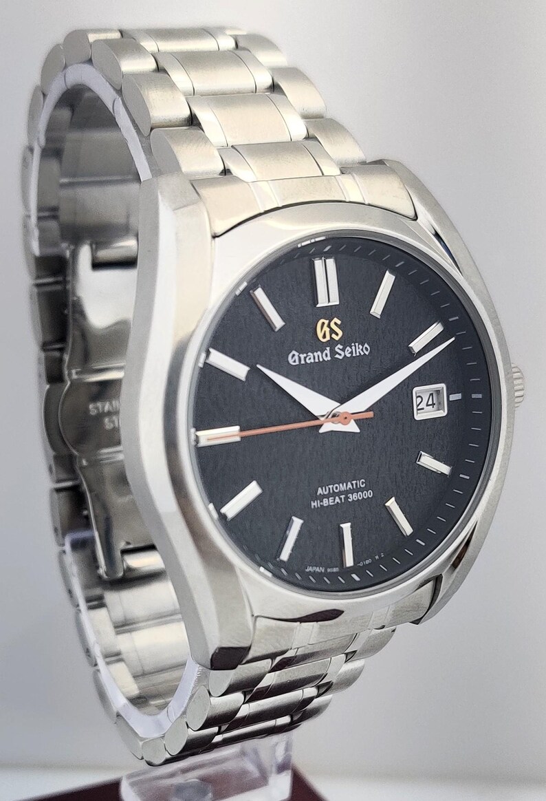 May include: A stainless steel Grand Seiko automatic wristwatch with a black dial. The watch features silver hands and markers, a date window, and the text "Grand Seiko" and "AUTOMATIC HI-BEAT 36000". The watch band is also stainless steel.