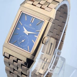 May include: A rose gold rectangular wristwatch with a blue dial. The watch features a small seconds subdial and gold hands. The watch band is also rose gold and has a woven design. The watch face reads "JAEGER-LECOULTRE".
