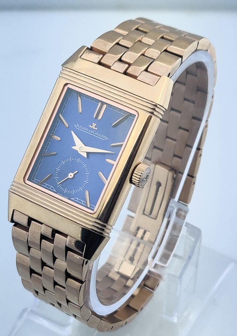 May include: A rose gold rectangular wristwatch with a blue dial. The watch features a small seconds subdial and gold hands. The watch band is also rose gold and has a woven design. The watch face reads "JAEGER-LECOULTRE".