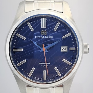 Könnte beinhalten: Eine Grand Seiko-Uhr aus Edelstahl mit blauem Zifferblatt und silbernen Zeigern und Markierungen. Das Zifferblatt zeigt das Datum und den Text "SPRING DRIVE 5 DAYS". Die Uhr hat ein silbernes Armband.