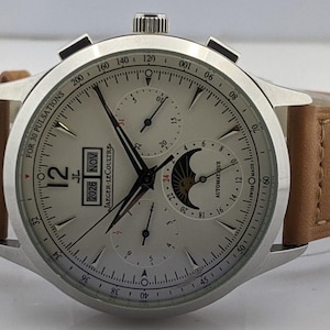 May include: A silver-toned wristwatch with a light-colored face and a brown leather strap. The watch face displays multiple sub-dials, hands, and the date. The text "JAEGER-LECOULTRE" is visible. The watch has a stainless steel case.
