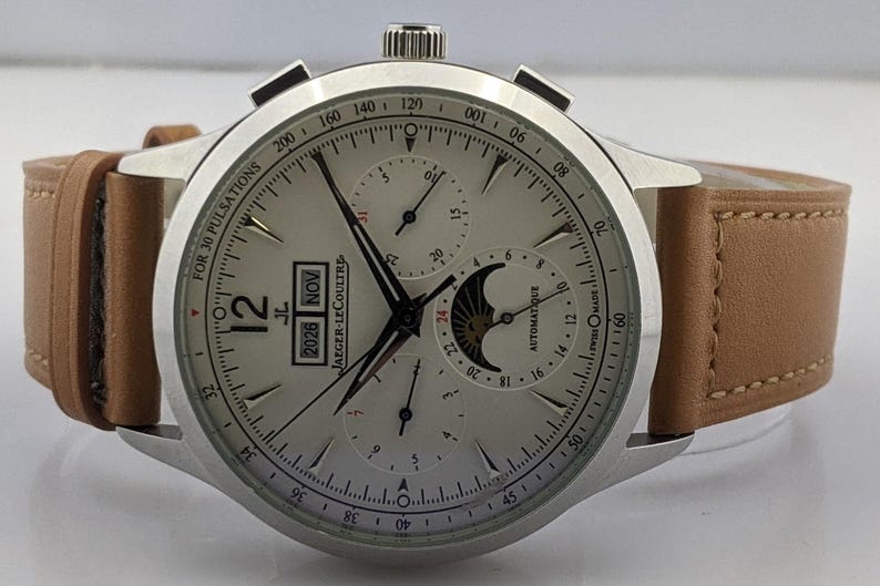 May include: A silver-toned wristwatch with a light-colored face and a brown leather strap. The watch face displays multiple sub-dials, hands, and the date. The text "JAEGER-LECOULTRE" is visible. The watch has a stainless steel case.