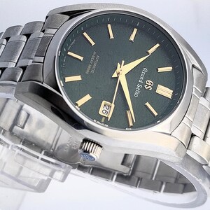 May include: A stainless steel Grand Seiko watch with a dark green dial and gold-colored hands and markers. The watch features a date display at the 3 o'clock position and a matching stainless steel bracelet. The watch is displayed on a clear stand.
