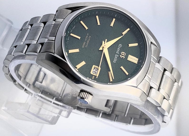May include: A stainless steel Grand Seiko watch with a dark green dial and gold-colored hands and markers. The watch features a date display at the 3 o'clock position and a matching stainless steel bracelet. The watch is displayed on a clear stand.