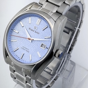 May include: A stainless steel wristwatch with a light blue textured dial. The watch features a date display at the 3 o'clock position and a silver-tone bracelet. The watch face has the words "Grand Seiko" printed on it.