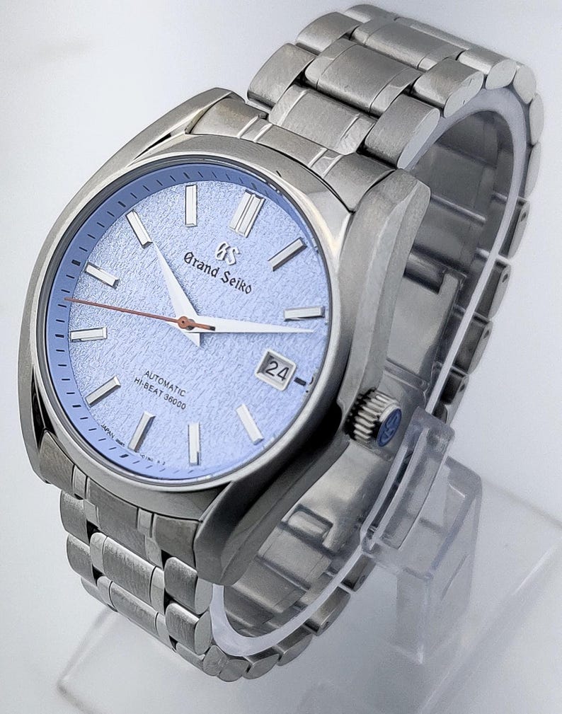 May include: A stainless steel wristwatch with a light blue textured dial. The watch features a date display at the 3 o'clock position and a silver-tone bracelet. The watch face has the words "Grand Seiko" printed on it.