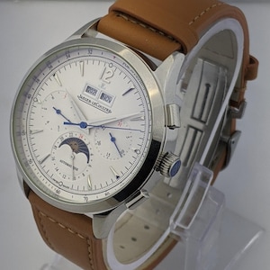 May include: A silver-toned wristwatch with a white face, featuring multiple subdials and a date display. The watch has a brown leather strap and the text "JAEGER-LECOULTRE" is visible on the face.