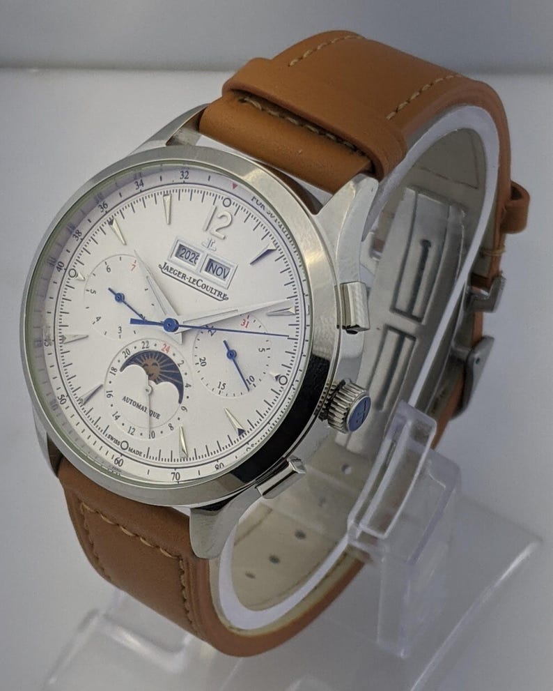 May include: A silver-toned wristwatch with a white face, featuring multiple subdials and a date display. The watch has a brown leather strap and the text "JAEGER-LECOULTRE" is visible on the face.