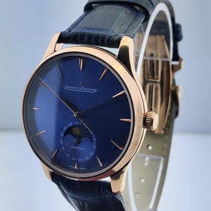 May include: A luxury wristwatch with a navy blue face and gold-toned casing. The watch features a leather band and the brand name "JAEGER-LECOULTRE" is visible. The watch has a subdial with a moon phase indicator and date display.