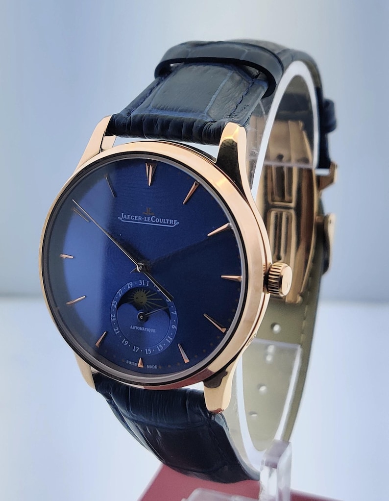 May include: A luxury wristwatch with a navy blue face and gold-toned casing. The watch features a leather band and the brand name "JAEGER-LECOULTRE" is visible. The watch has a subdial with a moon phase indicator and date display.