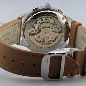 May include: Close-up of a silver wristwatch with a brown leather strap. The watch's back reveals intricate internal mechanisms. The strap is secured with a silver buckle. The watch is labeled "SWISS MADE 5 bar".