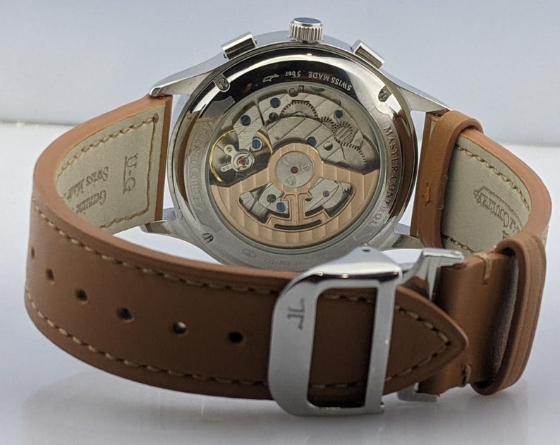 May include: Close-up of a silver wristwatch with a brown leather strap. The watch's back reveals intricate internal mechanisms. The strap is secured with a silver buckle. The watch is labeled "SWISS MADE 5 bar".