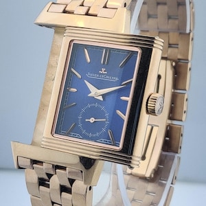 May include: A gold-toned wristwatch with a rectangular face and a blue dial. The watch features a small seconds subdial and gold hands. The band is made of gold-toned links. The watch is displayed on a clear stand.