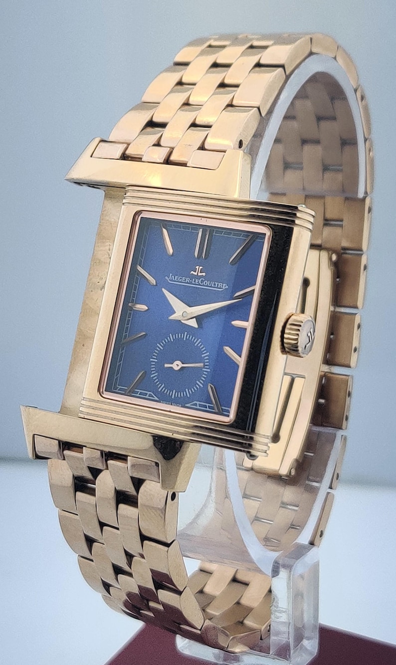 May include: A gold-toned wristwatch with a rectangular face and a blue dial. The watch features a small seconds subdial and gold hands. The band is made of gold-toned links. The watch is displayed on a clear stand.