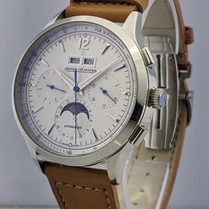 May include: A silver-toned wristwatch with a white face, featuring multiple dials and a moon phase indicator. The watch has a brown leather strap. The face displays the date as "2026 NOV" and the brand name "JAEGER-LECOULTRE".
