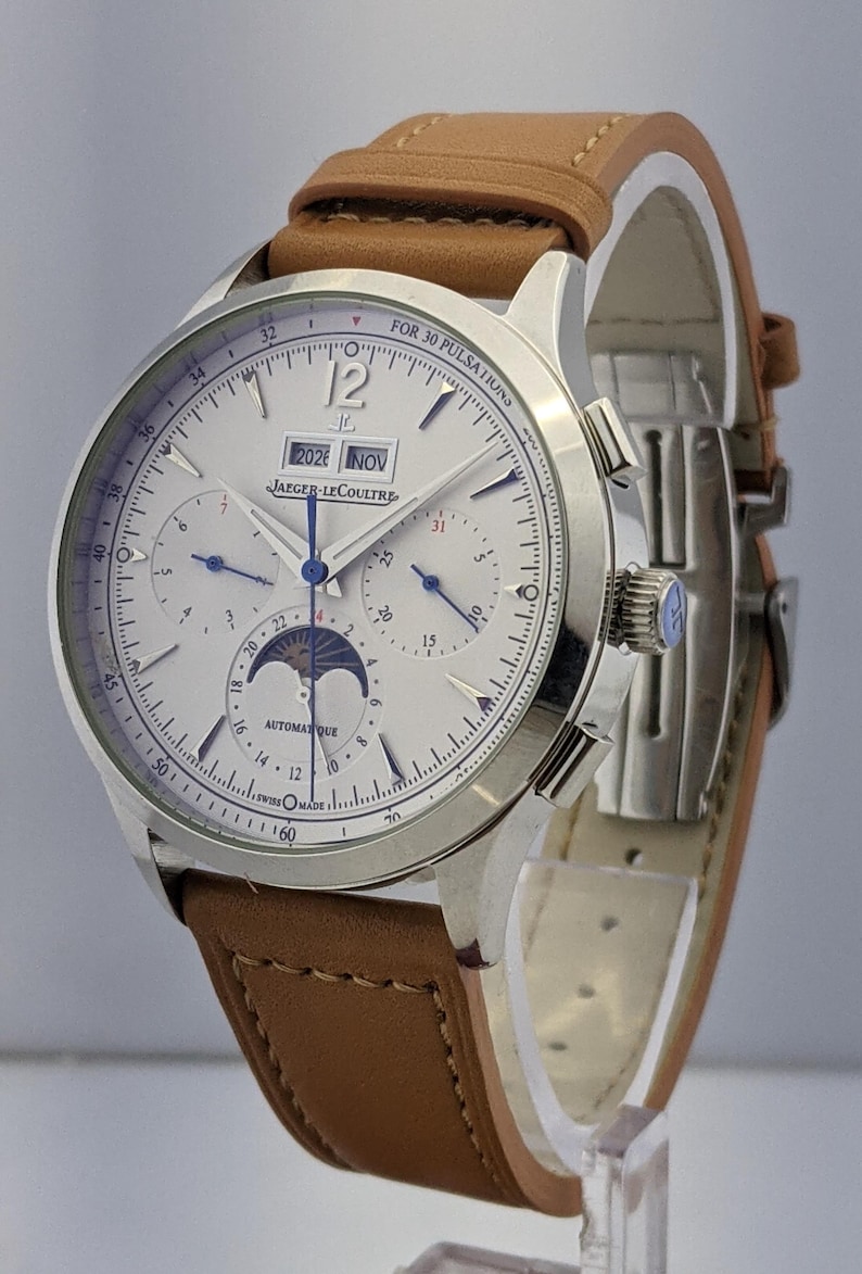 May include: A silver-toned wristwatch with a white face, featuring multiple dials and a moon phase indicator. The watch has a brown leather strap. The face displays the date as "2026 NOV" and the brand name "JAEGER-LECOULTRE".