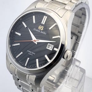 K&ouml;nnte beinhalten: Eine Grand Seiko Armbanduhr aus Edelstahl mit schwarzem Zifferblatt und silbernen Zeigern. Die Uhr verf&uuml;gt &uuml;ber eine Datumsanzeige bei 3 Uhr und ein silbernes Gliederarmband. Die Uhr wird auf einem transparenten St&auml;nder pr&auml;sentiert.