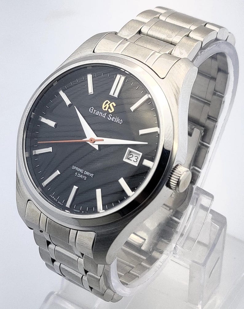 K&ouml;nnte beinhalten: Eine Grand Seiko Armbanduhr aus Edelstahl mit schwarzem Zifferblatt und silbernen Zeigern. Die Uhr verf&uuml;gt &uuml;ber eine Datumsanzeige bei 3 Uhr und ein silbernes Gliederarmband. Die Uhr wird auf einem transparenten St&auml;nder pr&auml;sentiert.