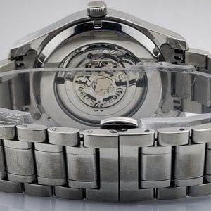 K&ouml;nnte beinhalten: Nahaufnahme einer silberfarbenen Armbanduhr, die das komplizierte mechanische Uhrwerk durch den transparenten R&uuml;cken zeigt. Die Uhr hat ein Edelstahlarmband mit einem sicheren Verschluss. Die Uhr ist neu und ungetragen.