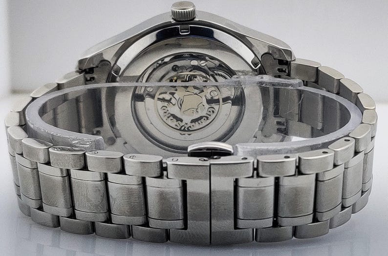 K&ouml;nnte beinhalten: Nahaufnahme einer silberfarbenen Armbanduhr, die das komplizierte mechanische Uhrwerk durch den transparenten R&uuml;cken zeigt. Die Uhr hat ein Edelstahlarmband mit einem sicheren Verschluss. Die Uhr ist neu und ungetragen.