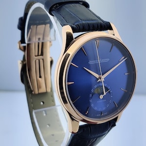 May include: A luxury wristwatch with a blue dial and gold-tone hands and case. The watch features a black leather strap and a deployment clasp. The watch face displays the Jaeger-LeCoultre logo.