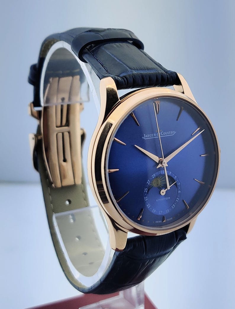 May include: A luxury wristwatch with a blue dial and gold-tone hands and case. The watch features a black leather strap and a deployment clasp. The watch face displays the Jaeger-LeCoultre logo.