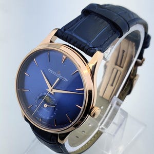 May include: A luxury wristwatch with a blue face and gold-tone hands and casing. The watch features a black leather band and the brand name "JAEGER-LECOULTRE" is visible on the watch face. The watch is displayed on a clear stand.