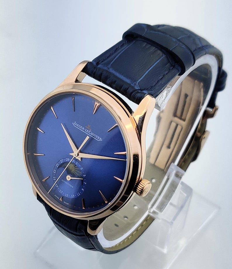 May include: A luxury wristwatch with a blue face and gold-tone hands and casing. The watch features a black leather band and the brand name "JAEGER-LECOULTRE" is visible on the watch face. The watch is displayed on a clear stand.