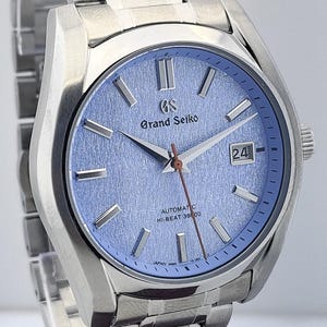 May include: A stainless steel wristwatch with a light blue textured dial. The watch features silver hands and markers, a date window, and the text "Grand Seiko" and "Automatic Hi-Beat 36000". The watch band is also stainless steel.