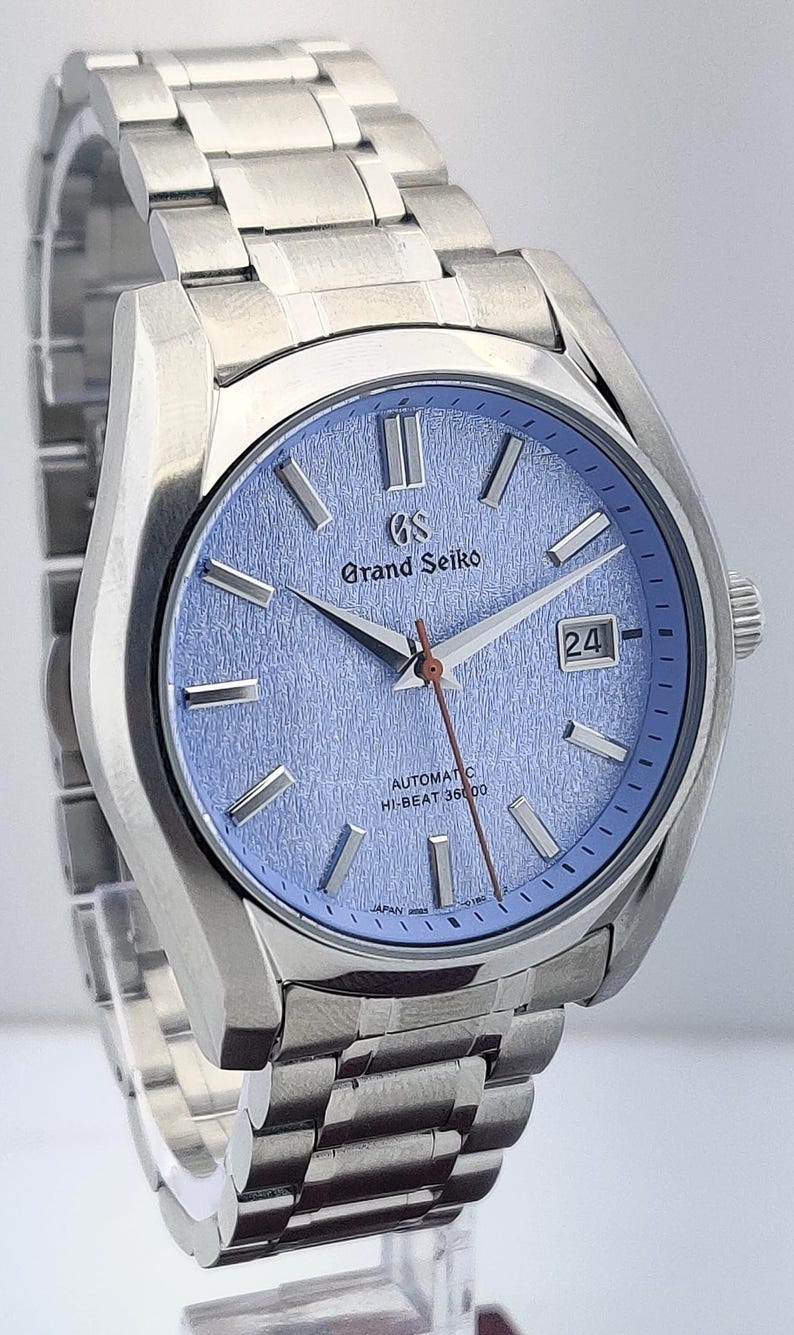 May include: A stainless steel wristwatch with a light blue textured dial. The watch features silver hands and markers, a date window, and the text "Grand Seiko" and "Automatic Hi-Beat 36000". The watch band is also stainless steel.